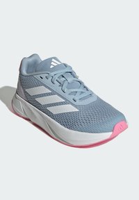 Zapatilla deportiva de color azul claro con parte superior de malla, franjas blancas y suela de goma rosa. Presenta punta redondeada y diseño con cordones.