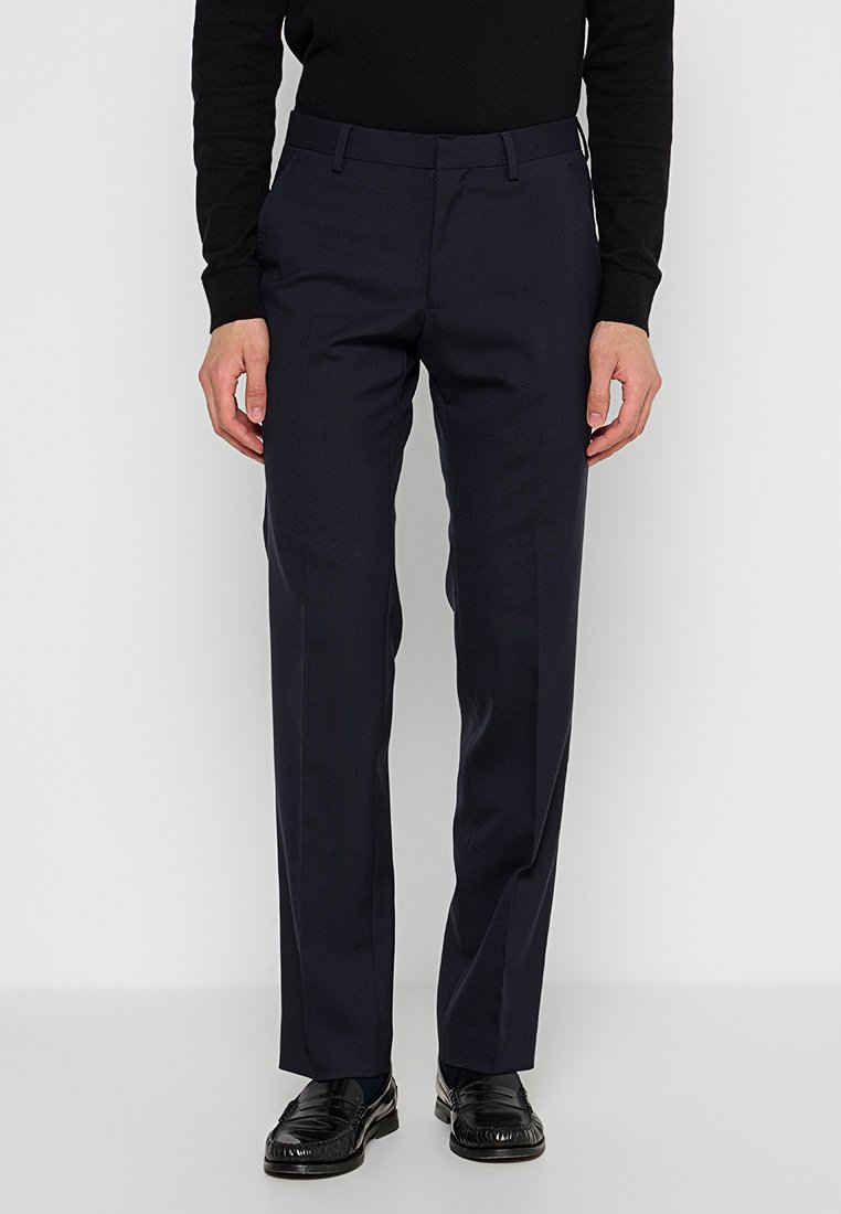 Pantalones de vestir azul marino fabricados en una tela suave, con un corte recto, con pasadores para el cinturón y bolsillos laterales. Llevados con zapatos negros y un suéter.