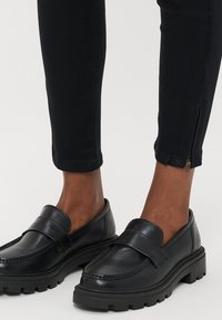 Črni usnjeni loaferji z debelim, nazobčanim podplatom. Imajo zasnovo za enostavno natikanje in širok trak preko zgornjega dela. Hlače so črne z zadrgami ob gležnjih.