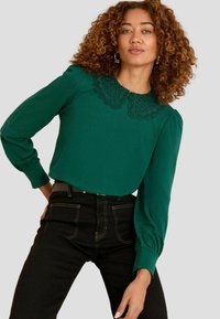 Grüne Bluse mit Spitzenverzierungen am Ausschnitt, langen puffärmeln, kombiniert mit hochtaillierten schwarzen Jeans. Einfaches und strukturiertes Design.