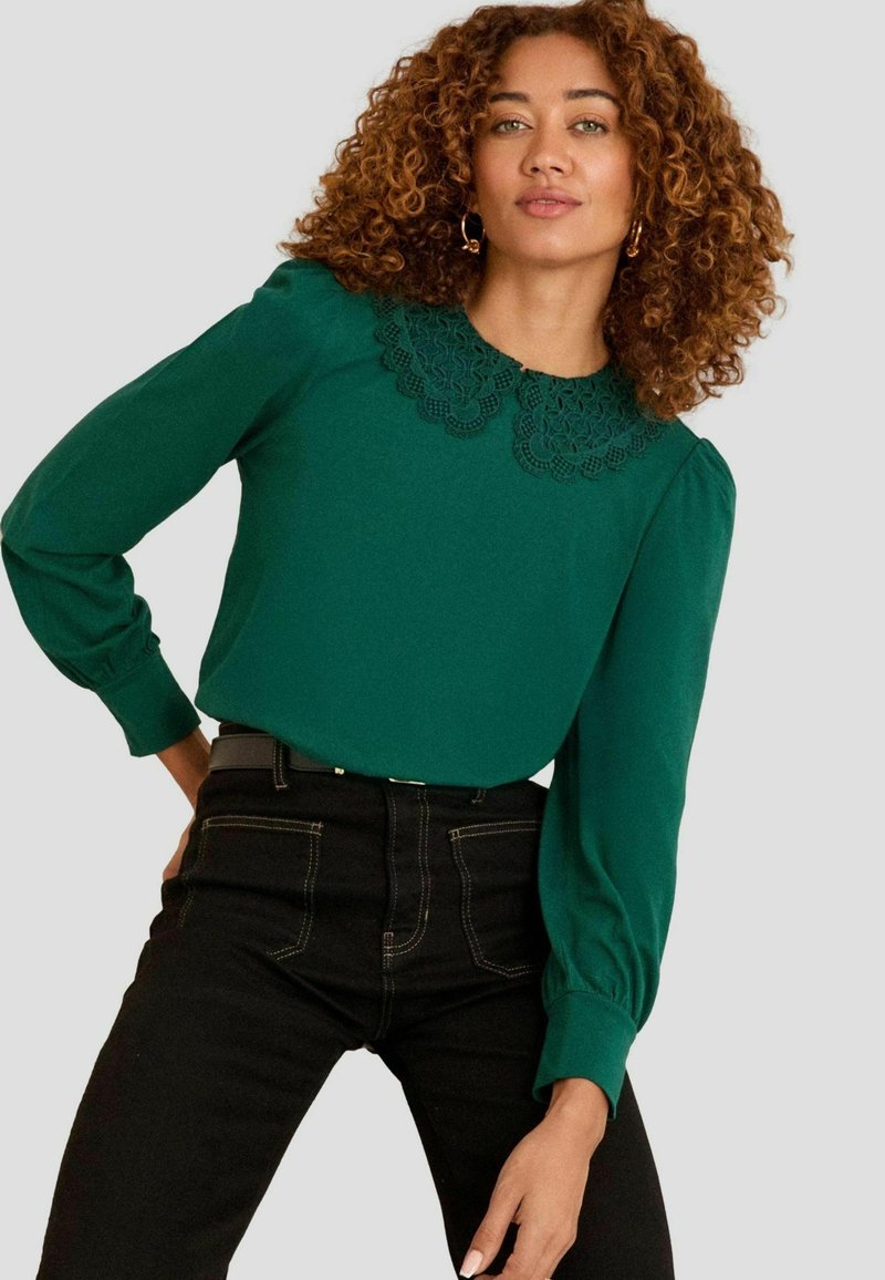 Grüne Bluse mit Spitzenverzierungen am Ausschnitt, langen puffärmeln, kombiniert mit hochtaillierten schwarzen Jeans. Einfaches und strukturiertes Design.