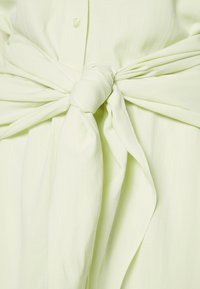 Gros plan d’un tissu jaune pâle noué à la taille, montrant la texture et un bouton au-dessus du nœud.