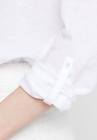 Bílá lněná košile s ohrnutými rukávy, vybavená uzávěrem na jeden knoflík. Měkká textura a lehká tkanina jsou viditelné v detailním pohledu.