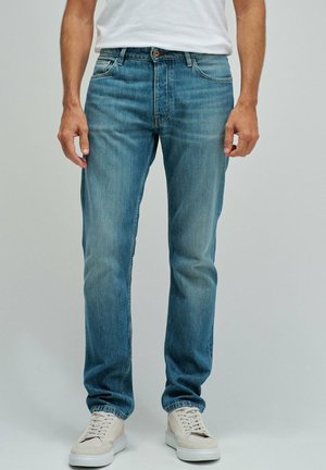 Hombre con vaqueros azul claro de corte recto, zapatillas blancas y camiseta blanca, de pie frente a un fondo liso y claro.