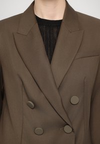 Bruine dubbele blazer met klepels, gemaakt van soepel materiaal, voorzien van ronde grijze knopen en een borstzak. Zwarte gestructureerde top eronder.