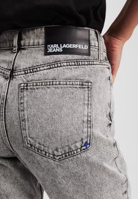 Jeans in denim grigio con un'etichetta in pelle nera visibile con la scritta "KARL LAGERFELD JEANS." Presentano un dettaglio accentuato blu sulla tasca posteriore.
