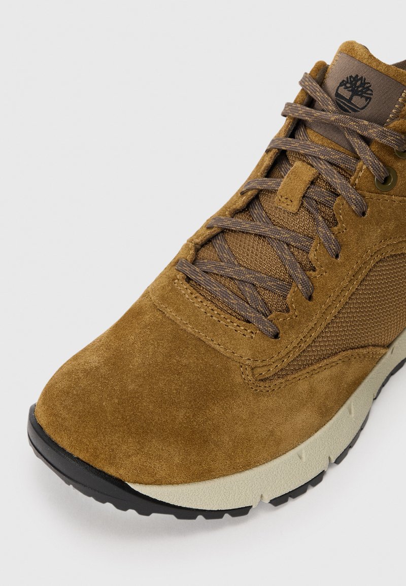 Zapatillas de gamuza marrón con acentos de malla, cordones texturizados y un logo en la lengua. Cuenta con una entresuela de color crema en contraste y una suela negra.