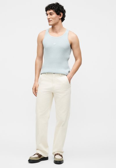 Νέος άνδρας στέκεται με τα χέρια στις τσέπες, φορώντας ανοιχτό μπλε ριπ αμάνικο, κρεμ παντελόνια και καφέ και λευκές μοκασίνες.