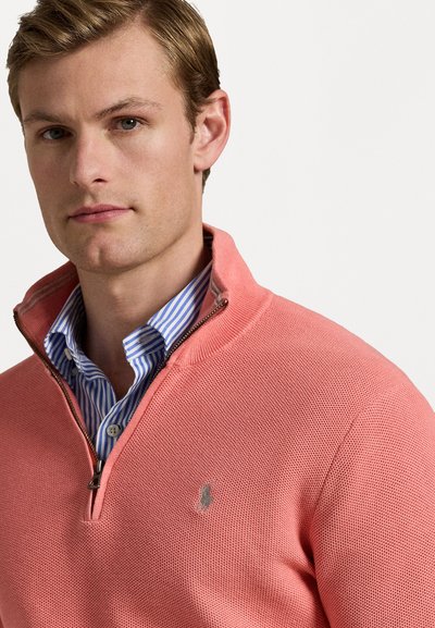 Un gilet zippé de couleur corail clair avec un tricot texturé, doté d'un col haut et d'un petit logo. Porté par-dessus une chemise rayée bleue et blanche.