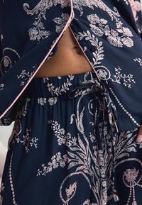 Jūrinės audinės rinkinys su rožiniais gėlės raštais; turi laisvą prigludimą viršuje su mygtukais ir derančias laisvas šortus su elastiniu juosmeniu ir raišteliu.