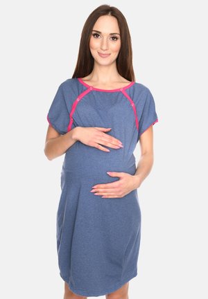 Schwangere Frau in einem blauen Kleid mit pinkem Saum, die sanft ihren Bauch hält, vor einem schlichten weißen Hintergrund stehend.