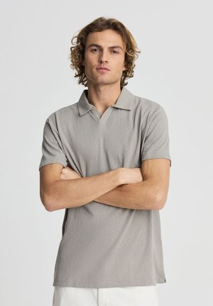 Giovane uomo con capelli biondi ricci che indossa una polo grigio chiaro a maniche corte, in piedi con le braccia incrociate davanti a uno sfondo semplice.