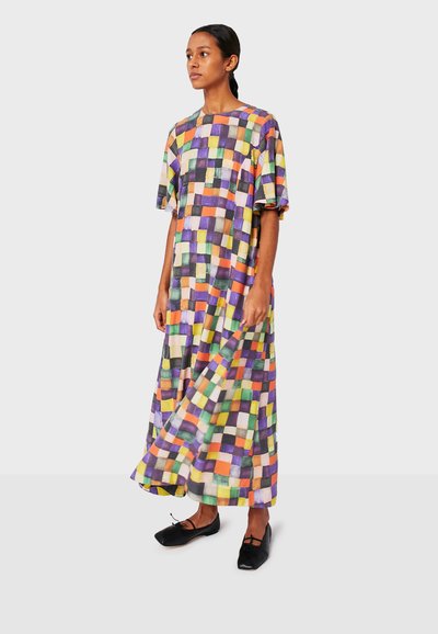 Donna in un maxi abito a scacchi colorato con maniche corte, che indossa ballerine nere su uno sfondo neutro.