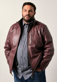 Burgundská kožená bunda s kapsou na zip, stojáčkem a hladkým povrchem, nošená přes šedou košili s knoflíky. Viditelné modré džíny pod tím.