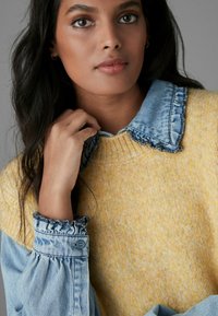 Pull en maille jaune avec un col en denim à volants, présentant de longues manches en denim. Texture légère, design moderne en couches et détail de bouton sur les poignets.