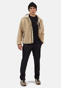 Beige fleecová bunda s černými akcenty, s kapsami na zip, v kombinaci s černými kalhotami a béžovými botami se černými pruhy. Černý pletený kulich.