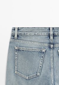 Vue arrière d'un jean en denim bleu clair montrant la partie supérieure de la taille, les passants de ceinture et la poche arrière gauche avec des coutures visibles et la texture du tissu.