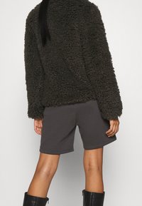 Veste marron en peluche avec une texture douce et des manches longues, associée à un short gris. Des bottes noires montantes complètent la tenue, offrant un look décontracté.