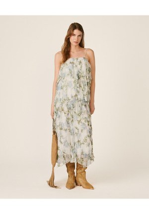 Mujer con un vestido maxi blanco con flores y tirantes finos, sosteniendo un bolso marrón con flecos, llevando botas de vaquero marrones, de pie sobre un fondo liso.