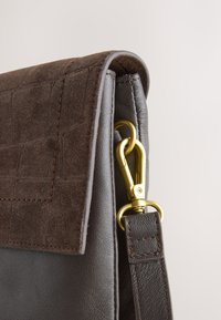 Gros plan sur un sac en cuir marron avec un rabat texturé et une boucle en métal doré attachant une sangle sur le côté.