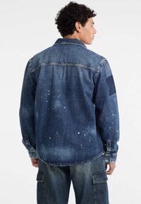 Donkerblauwe denimjack met spetters verf, een klassieke kraag en knopenmanchet. Heeft een patch op de linker schouder.