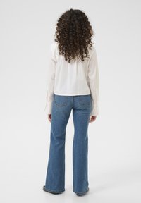 White, collared, long-sleeve shirt with a straight cut; paired with flared, medium-blue jeans. Curly hair falls to shoulders.