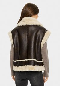 Ruskea shearling-liivi, jossa on vaalea shearling-kaulus ja helma, istuva malli, hihaton, teksturoitu pinta ja näkyvät saumat.