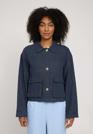 Woman with curly hair wearing a dark blue buttoned jacket with front pockets and light blue pants, standing against a plain background.