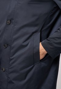 Marineblå vandtæt frakke med knaplukning, sidelommer og glat stofstruktur. Arm synlig med hånd i lommen.