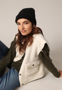 Fleece vest in crèmekleur met voorzakken, gedragen over een donkergroene trui. Zwarte gebreide beanie. Casual spijkerbroek. Neutrale achtergrond.