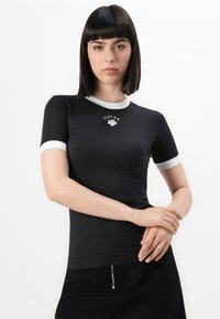 Svart tettsittende t-skjorte med hvit ribbet krage og ermer, med "GUESS" logo i sølv på forsiden. Mykt materiale, korte ermer.