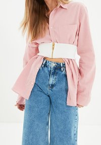 Nainen, jolla on yllään löysä vaaleanpunainen napitettava paita, leveä valkoinen vetoketju korsetti vyö ja korkeavyötäröiset leveät siniset farkut.