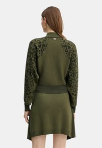 Donna che indossa un abito a maglia verde oliva con macchie leopardate nere sulle maniche e sulla parte superiore della schiena, in piedi con le spalle rivolte verso la fotocamera.
