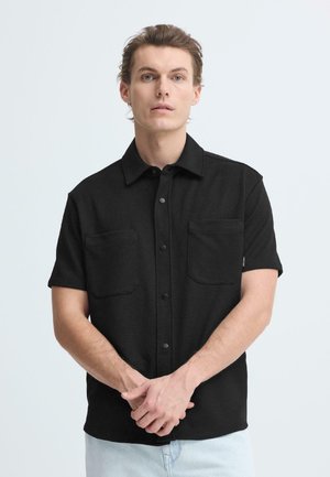 Sort kortærmet skjorte lavet af tekstureret stof med to forlommer og knaplukning, båret med lyseblå jeans.