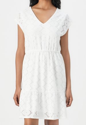 Vrouw met kort mouwtje, witte eyelet jurk met V-hals en elastische taille, staand tegen een effen witte achtergrond.