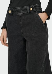 Črne široke denim hlače z visokim pasom, ki imajo dva zlata gumba, stranske žepke in rahlo teksturirano površino.