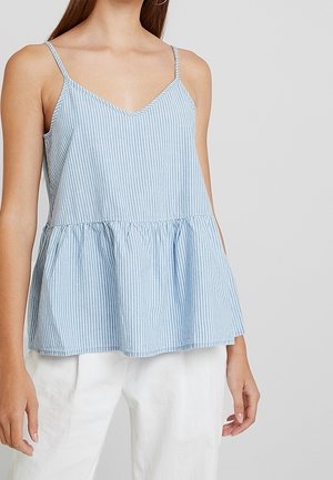 Femme portant un top sans manches à rayures verticales bleu clair et blanc avec de fines bretelles et un pantalon blanc sur un fond gris uni.