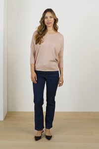 Svetlo roza prevelik pulover s tremi četrtinskimi rokavi, v kombinaciji z temno modrimi ravnimi hlačami in črnimi salonarji na leseni tal.