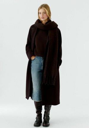 Un long manteau marron foncé, un haut côtelé sombre, une jupe en denim, des bottes marron jusqu'aux genoux et une écharpe assortie foncée avec des franges.