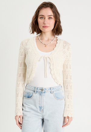 Joven con cabello corto y castaño vistiendo un cárdigan blanco de crochet sobre una camiseta blanca y jeans de tiro alto azul claro contra un fondo liso.