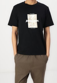 Osoba ubrana w czarną koszulkę z beżowym abstrakcyjnym nadrukiem i białym tekstem, w połączeniu z brązowymi spodniami, z rękami w kieszeniach na białym tle.