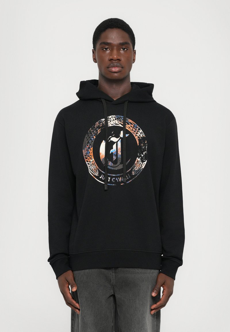 Joven vistiendo una sudadera negra con capucha que tiene un gráfico circular multicolor y el texto "Just Cavalli" en el pecho, combinada con unos jeans grises.