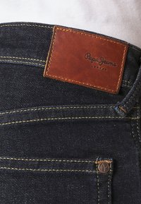 Jeans i denim med en mörk indigo färg, som har gula sömmar, en brun läderetikett med "Pepe Jeans London" och en liten metall-logotyp på fickan.