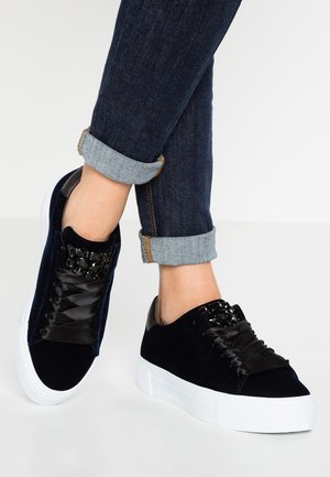 Osoba nosząca podwinięte ciemnoniebieskie dżinsy i czarne aksamitne trampki na platformie z grubymi białymi podeszwami oraz czarnymi satynowymi sznurowadłami.