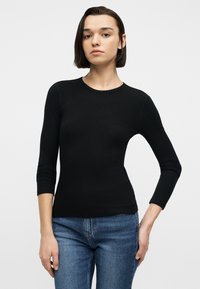 Νεαρό άτομο με κοντά σκούρα μαλλιά που φορά εφαρμοστή μαύρη μπλούζα με μακριά μανίκια και μπλε τζιν, στέκεται μπροστά σε απλό λευκό φόντο.