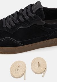 Black suede sneaker with dark grey laces, rubber outsole in brown. Two cream-colored laces rolled beside the shoe. Accents of black leather.