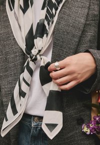 Schwarz-weiß gemusterter Schal aus leichtem Stoff, zu einem Knoten gebunden, über einem grauen Blazer getragen, mit einem silbernen Ring, der an der Hand sichtbar ist.
