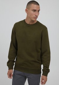 Oliven grønn genser i et mykt stoff, med rund hals, ribbede ermer, og en liten logo på brystet, kombinert med grå bukser.