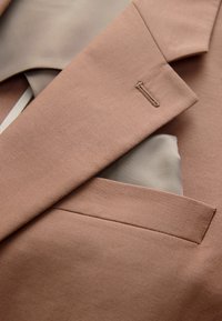 Nahaufnahme eines hellbraunen Sakko-Revers und einer Tasche mit einem beige gefalteten Einstecktuch.