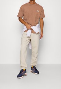 Mann in einem braunen T-Shirt, beigen Jogginghosen, marineblauen Turnschuhen und einem hellgrauen Sweatshirt, das um die Taille gebunden ist, steht vor einem schlichten Hintergrund.